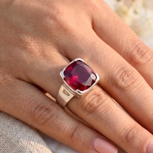 May include: A silver ring featuring a large, square-cut, deep red gemstone. The ring has a thick band and a simple, modern design. The gemstone is the focal point, catching the light and creating a bold statement. The ring is shown on a hand.