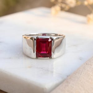 May include: A silver ring featuring a rectangular, deep red gemstone. The ring has a wide band and a polished finish, set on a white marble surface. The gemstone is the focal point, displaying a vibrant colour.