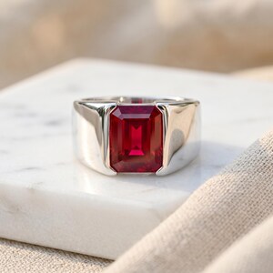 Puede incluir: Un anillo de plata con una gran gema rectangular de color rojo intenso. El anillo tiene una banda ancha y un acabado pulido, que resalta el color vibrante de la piedra. El anillo está sobre una superficie blanca.