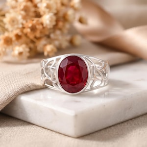 Puede incluir: Un anillo de plata con una gema ovalada de color rojo intenso. El anillo tiene un diseño calado alrededor de la gema. El anillo se muestra sobre una superficie de mármol blanco.