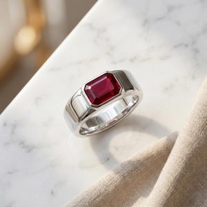 May include: A silver ring with a rectangular, deep red gemstone. The ring has a polished, smooth band and a bezel setting that frames the gemstone. The ring is displayed on a white marble surface.