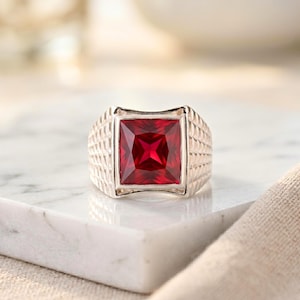 May include: A silver ring featuring a large, square, red gemstone. The ring has a textured band and a polished finish. The gemstone is a deep red colour, and the ring is set on a white surface.