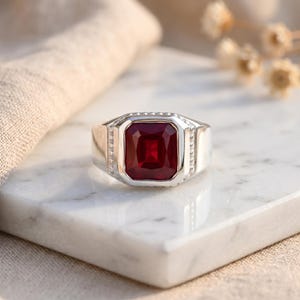 May include: A silver ring with a large, rectangular, deep red gemstone. The ring has a wide band with a geometric design and small, clear accent stones. The gemstone is the focal point, set in a polished silver bezel.