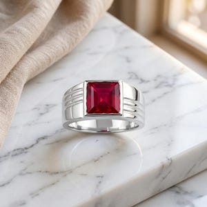 Puede incluir: Un anillo plateado con una gran gema roja rubí cuadrada. El anillo tiene una banda ancha con ranuras horizontales en los lados. La gema está engastada en un bisel cuadrado, creando un diseño audaz. El anillo se exhibe sobre una superficie de mármol.