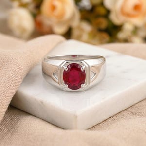 May include: A silver ring featuring a large, oval-cut ruby gemstone. The ruby is a deep red hue, set in a rectangular bezel. The ring has a simple, classic design.