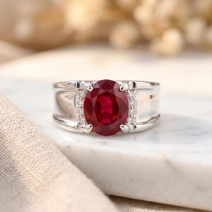 Peut inclure: Une bague en argent ornée d'un grand rubis ovale. Le rubis est d'un rouge profond et constitue le point central de la bague. De petites pierres précieuses claires sont serties de chaque côté du rubis, ajoutant de l'éclat. La bague est posée sur une surface blanche.