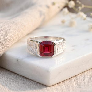 May include: A silver ring with a large, rectangular, deep red gemstone. The ring has a floral pattern engraved on the sides. The ring is displayed on a white marble surface.