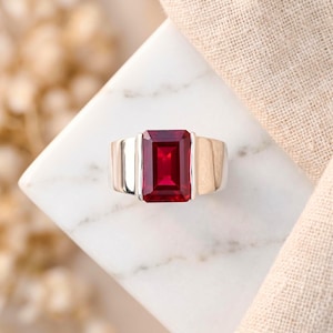 Könnte beinhalten: Ein silberner Ring mit einem großen, rechteckigen, tiefroten Edelstein. Der Ring hat ein modernes Design mit einem breiten Band und einer prominenten Fassung für den Edelstein. Der Ring wird auf einer weißen Marmoroberfläche präsentiert.