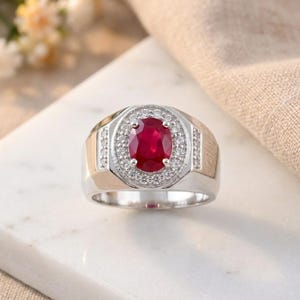 May include: A silver ring featuring a large oval ruby gemstone at the centre, encircled by a halo of smaller, clear gemstones. The ring has a wide band with gold-coloured accents on the sides.