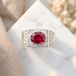 May include: A silver ring with a large, oval-cut, deep red gemstone at the center. The ring has a detailed, textured design with small, clear accent stones. The ring is displayed on a white surface.