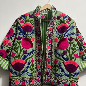 Peut inclure: Veste vert clair ornée d'un motif floral. La veste présente de grandes fleurs roses et violettes avec des feuilles bleues et vertes. Les poignets sont retroussés, révélant une doublure rayée verte et blanche. La veste est suspendue à un cintre en bois.