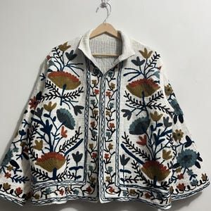 Peut inclure: Veste blanche avec broderies florales colorées. La veste a un col montant et une fermeture boutonnée. La broderie comprend des nuances d'orange, de vert, de bleu et de marron. La veste est suspendue à un cintre en bois.