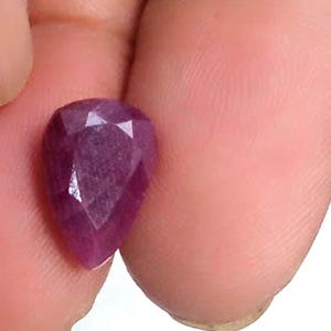 Könnte beinhalten: Ein birnenförmiger, facettierter Edelstein in einem tiefen Magenta-Violett-Ton. Der Stein hat eine spitz zulaufende Unterseite und eine abgerundete Oberseite, mit zahlreichen dreieckigen Facetten auf der Oberfläche, die das Licht reflektieren und ein strukturiertes, kristallines Aussehen erzeugen.