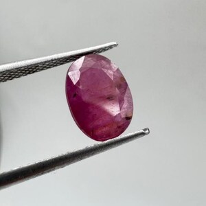Könnte beinhalten: Ein facettierter, oval geformter Edelstein mit einem tiefen rosa-roten Farbton, der von einer silberfarbenen Metallpinzette gehalten wird. Der Stein zeigt eine durchscheinende Qualität mit sichtbaren Einschlüssen und einer polierten Oberfläche.