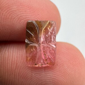Könnte beinhalten: Ein rechteckiger, durchscheinender Edelstein mit einem Farbverlauf von Rosa bis Goldbraun. Die Oberfläche des Steins ist mit einem symmetrischen, blattartigen Muster versehen, das aus tiefen, linearen Rillen besteht, die das Licht reflektieren.