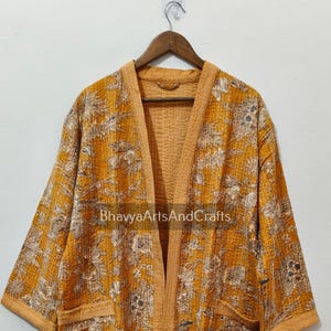 May include: Mustard yellow Kantha robe with floral and leaf patterns. The robe has long sleeves, two front pockets, and the text "BhavyaArtsAndCrafts". This lightweight, open-front robe is perfect for layering.
