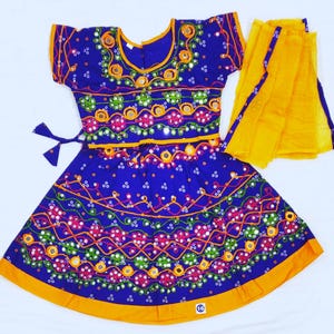 May include: A vibrant blue traditional Indian dress with intricate embroidery and mirror work. The dress features a short-sleeved top and a flared skirt, both adorned with colorful patterns. A bright yellow scarf is draped over the dress, adding a contrasting element.