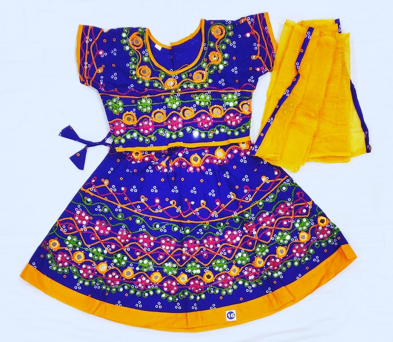 May include: A vibrant blue traditional Indian dress with intricate embroidery and mirror work. The dress features a short-sleeved top and a flared skirt, both adorned with colorful patterns. A bright yellow scarf is draped over the dress, adding a contrasting element.