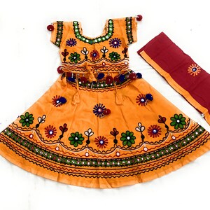 May include: An orange traditional outfit with intricate embroidery and mirror work. The set includes a short-sleeved top and a flared skirt, both adorned with floral patterns in green, red, and blue. A matching red scarf with embroidery completes the ensemble.