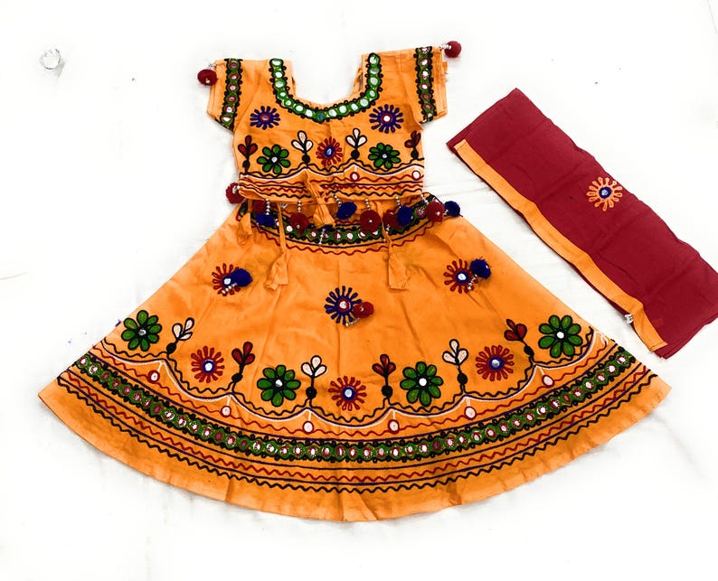May include: An orange traditional outfit with intricate embroidery and mirror work. The set includes a short-sleeved top and a flared skirt, both adorned with floral patterns in green, red, and blue. A matching red scarf with embroidery completes the ensemble.