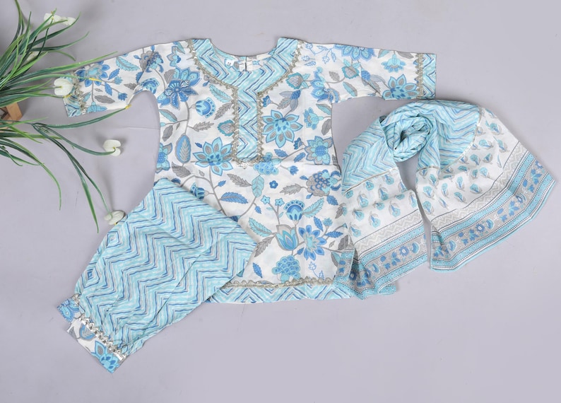 K&ouml;nnte beinhalten: Ein wei&szlig;-blaues Outfit mit Blumenmuster, bestehend aus einem Oberteil, einer Hose und einem Schal. Das Oberteil hat einen runden Ausschnitt und lange &Auml;rmel. Die Hose hat ein Zickzackmuster. Der Schal hat ein Blumen- und Paisley-Design.
