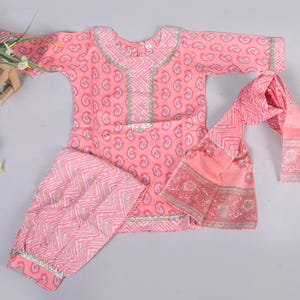 Puede incluir: Un conjunto rosa con estampado de cachemira para niños, que incluye una parte superior de manga larga, pantalones y una bufanda. El conjunto presenta ribetes plateados y un patrón geométrico a juego en los pantalones y la bufanda. La ropa está sobre una superficie gris claro.