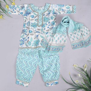 K&ouml;nnte beinhalten: Ein Kinder-Outfit mit wei&szlig;em und blauem Blumenmuster, bestehend aus einem lang&auml;rmeligen Oberteil, einer Hose und einem Schal. Das Oberteil und der Schal zeigen ein Blumenmuster, w&auml;hrend die Hose ein Zickzack-Design hat. Das Outfit ist mit goldenen Details verziert.