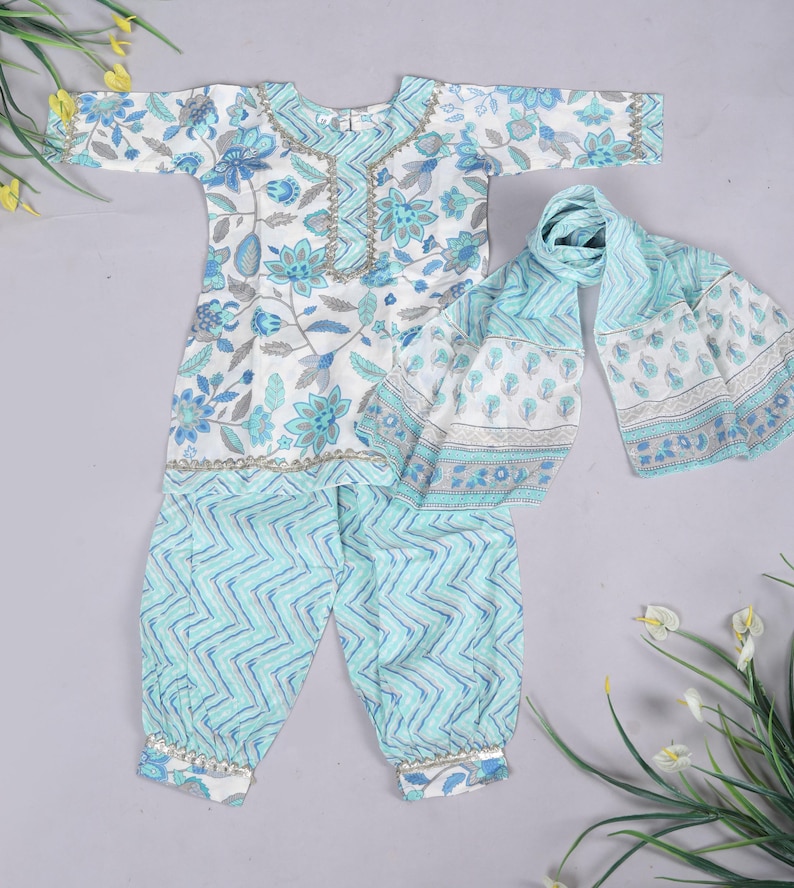 K&ouml;nnte beinhalten: Ein Kinder-Outfit mit wei&szlig;em und blauem Blumenmuster, bestehend aus einem lang&auml;rmeligen Oberteil, einer Hose und einem Schal. Das Oberteil und der Schal zeigen ein Blumenmuster, w&auml;hrend die Hose ein Zickzack-Design hat. Das Outfit ist mit goldenen Details verziert.