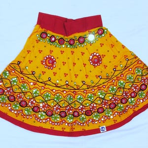May include: A vibrant yellow skirt with intricate embroidery and mirror work. The skirt features a red waistband and hem, with detailed patterns in red, green, and white. The design includes floral and geometric motifs, creating a festive look.