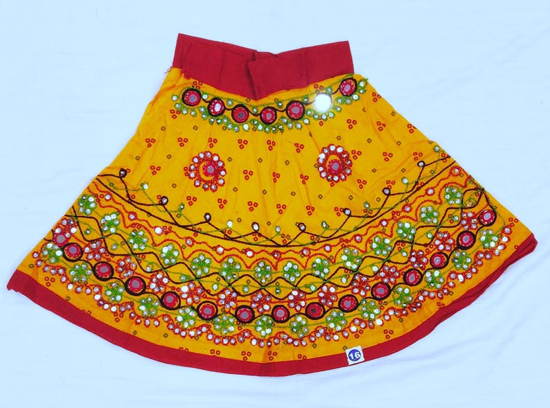May include: A vibrant yellow skirt with intricate embroidery and mirror work. The skirt features a red waistband and hem, with detailed patterns in red, green, and white. The design includes floral and geometric motifs, creating a festive look.
