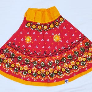 May include: A red skirt with yellow trim and a yellow waistband. The skirt is decorated with embroidered floral patterns, mirrors, and white dots. The skirt is a vibrant piece of clothing.