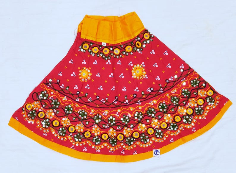 May include: A red skirt with yellow trim and a yellow waistband. The skirt is decorated with embroidered floral patterns, mirrors, and white dots. The skirt is a vibrant piece of clothing.