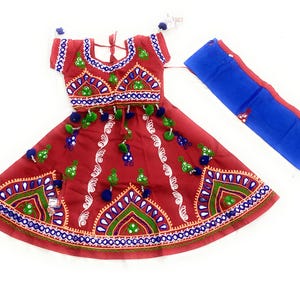 May include: A red traditional Indian outfit with intricate embroidery in green, blue, and white. The outfit includes a top, skirt, and a blue scarf. The skirt and top are adorned with decorative tassels.