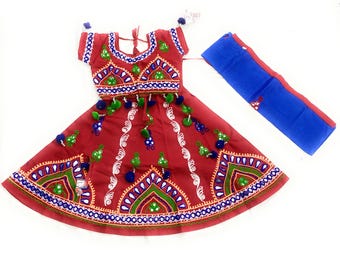 May include: A red traditional Indian outfit with intricate embroidery in green, blue, and white. The outfit includes a top, skirt, and a blue scarf. The skirt and top are adorned with decorative tassels.