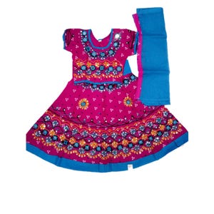 May include: A magenta-colored traditional Indian dress with intricate embroidery and mirror work. The dress features a short-sleeved top and a flared skirt, both adorned with blue and gold accents. A matching blue scarf is included.