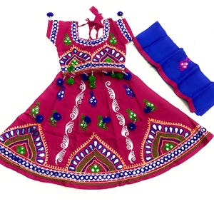 May include: A magenta-colored traditional Indian outfit, featuring a short-sleeved top and a flared skirt, both adorned with intricate embroidery in white, blue, green, and gold. A matching blue scarf with pink trim is included. The outfit is decorated with small pom-poms.