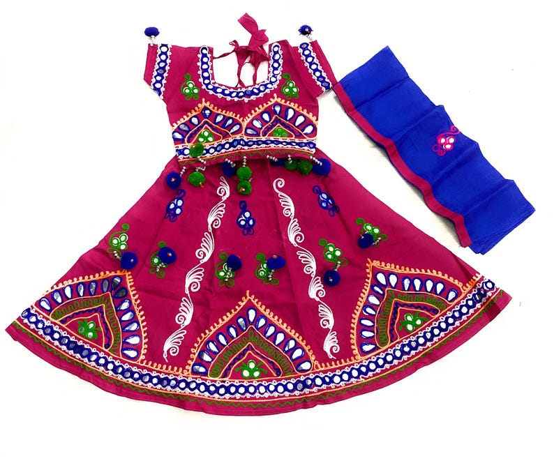 May include: A magenta-colored traditional Indian outfit, featuring a short-sleeved top and a flared skirt, both adorned with intricate embroidery in white, blue, green, and gold. A matching blue scarf with pink trim is included. The outfit is decorated with small pom-poms.