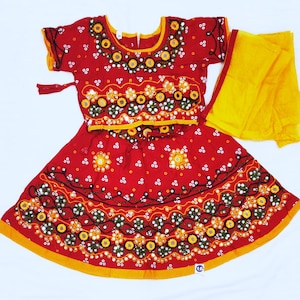 May include: A red traditional Indian dress with intricate embroidery and mirror work. The dress features a short-sleeved top and a flared skirt, both adorned with floral and geometric patterns. A yellow scarf is draped over the shoulder.