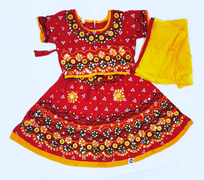 May include: A red traditional Indian dress with intricate embroidery and mirror work. The dress features a short-sleeved top and a flared skirt, both adorned with floral and geometric patterns. A yellow scarf is draped over the shoulder.