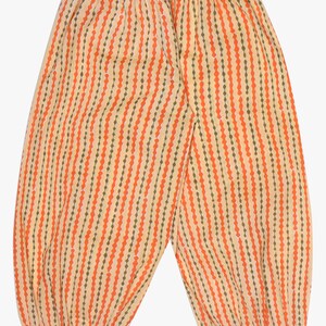 K&ouml;nnte beinhalten: Eine Hose mit orangefarbenem und gelbem Muster mit vertikalen Streifen. Die Hose hat einen gerafften Bund und Manschetten mit floralen und silbernen Verzierungen. Der Stoff hat ein gepunktetes Streifendesign in Orange und Olivgr&uuml;n.