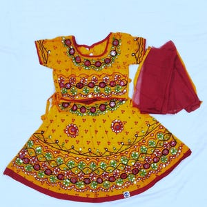 Puede incluir: Un conjunto tradicional amarillo con ribetes rojos y bordados intrincados. El conjunto incluye una parte superior de manga corta, una falda y una bufanda roja. El atuendo presenta patrones detallados con espejos y acentos coloridos.