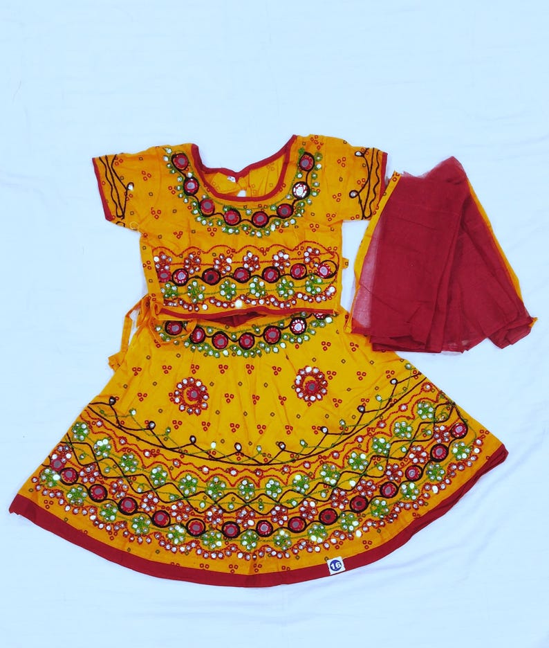May include: A yellow traditional outfit with red trim and intricate embroidery. The set includes a short-sleeved top, a skirt, and a red scarf. The outfit features detailed patterns with mirrors and colorful accents, showcasing a cultural design.