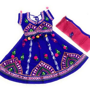 May include: A vibrant blue traditional Indian outfit, featuring intricate embroidery with pink, green, and white accents. The set includes a top, skirt, and a pink scarf. The skirt and top are adorned with mirrors and pom-poms.