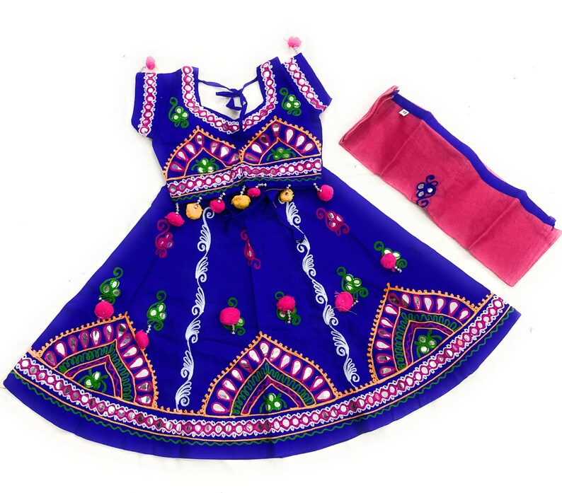 May include: A vibrant blue traditional Indian outfit, featuring intricate embroidery with pink, green, and white accents. The set includes a top, skirt, and a pink scarf. The skirt and top are adorned with mirrors and pom-poms.