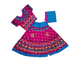 May include: A three-piece magenta and blue traditional outfit. The set includes a short-sleeved top, a skirt, and a scarf. The garments are adorned with intricate patterns and embellishments in shades of blue, orange, and gold. The skirt and top have a blue trim.