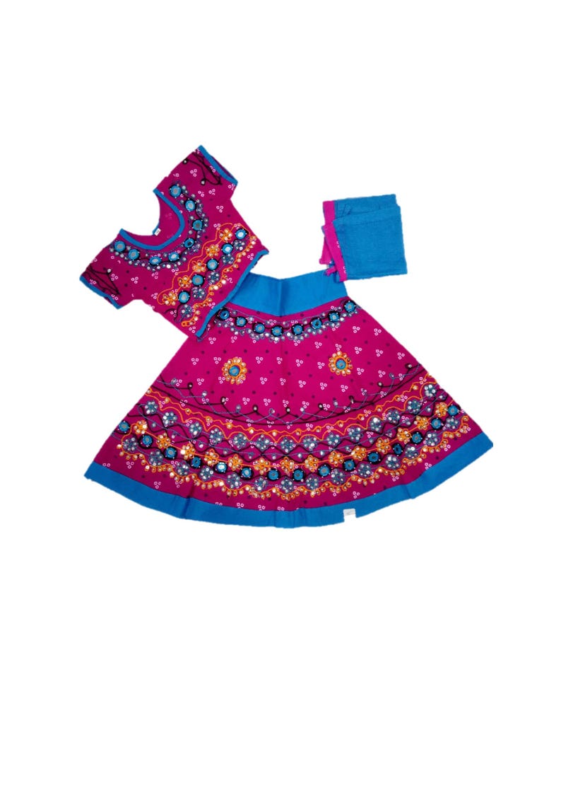 May include: A three-piece magenta and blue traditional outfit. The set includes a short-sleeved top, a skirt, and a scarf. The garments are adorned with intricate patterns and embellishments in shades of blue, orange, and gold. The skirt and top have a blue trim.