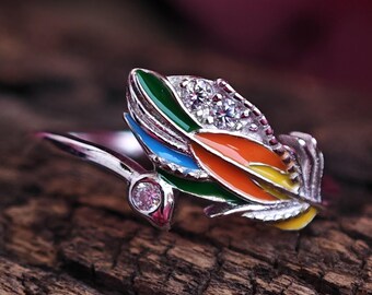 Puede incluir: Anillo plateado con un diseño de pluma colorida. La pluma tiene secciones de esmalte verde, azul, naranja y amarillo, con cristales transparentes y una pequeña gema rosa. El anillo está sobre una superficie de madera texturizada.