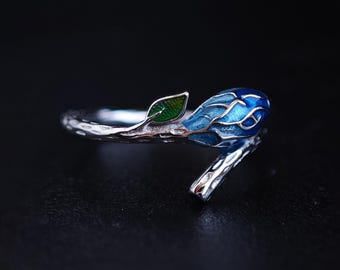 Puede incluir: Anillo plateado con un diseño de pájaro azul y plateado con un acento de hoja verde. El pájaro tiene detalles intrincados de esmalte azul. El anillo se encuentra sobre un fondo oscuro, resaltando los detalles de la joya.