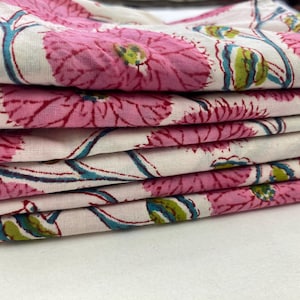 Op de afbeelding: Een stapel gevouwen stof met een bloemenpatroon. Het ontwerp toont grote roze bloemen met groene en blauwe accenten op een witte achtergrond. De stof lijkt katoen of een vergelijkbaar lichtgewicht materiaal te zijn.