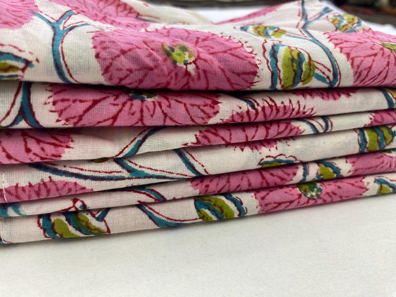 Op de afbeelding: Een stapel gevouwen stof met een bloemenpatroon. Het ontwerp toont grote roze bloemen met groene en blauwe accenten op een witte achtergrond. De stof lijkt katoen of een vergelijkbaar lichtgewicht materiaal te zijn.